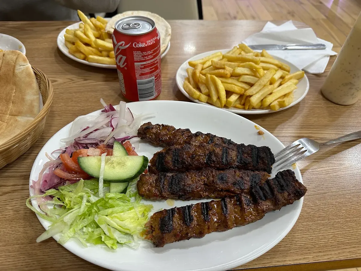 Four charcoal-grilled Adana kebab skewers on a plate with red onion, tomato, cucumber and lettuce, with chips and bread